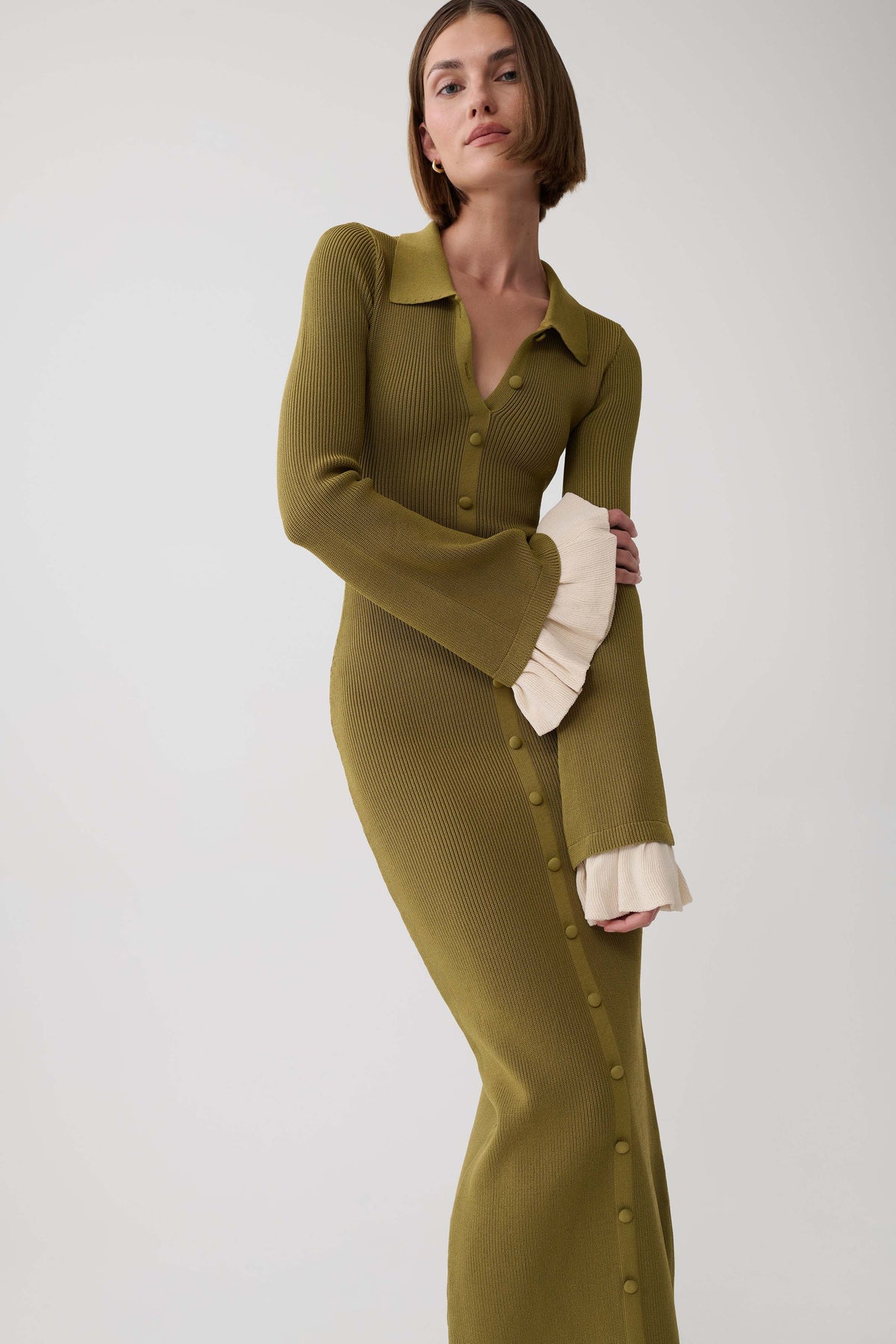 A woman models a ribbed olive green long-sleeve collared dress with cream ruffled cuffs and a button front detail, set against a plain light gray studio background.