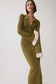 A woman models a ribbed olive green long-sleeve collared dress with cream ruffled cuffs and a button front detail, set against a plain light gray studio background.
