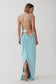 A woman modeling a light blue maxi dress from the back, showing an open back with thin straps and a draped skirt, against a plain light gray studio background.