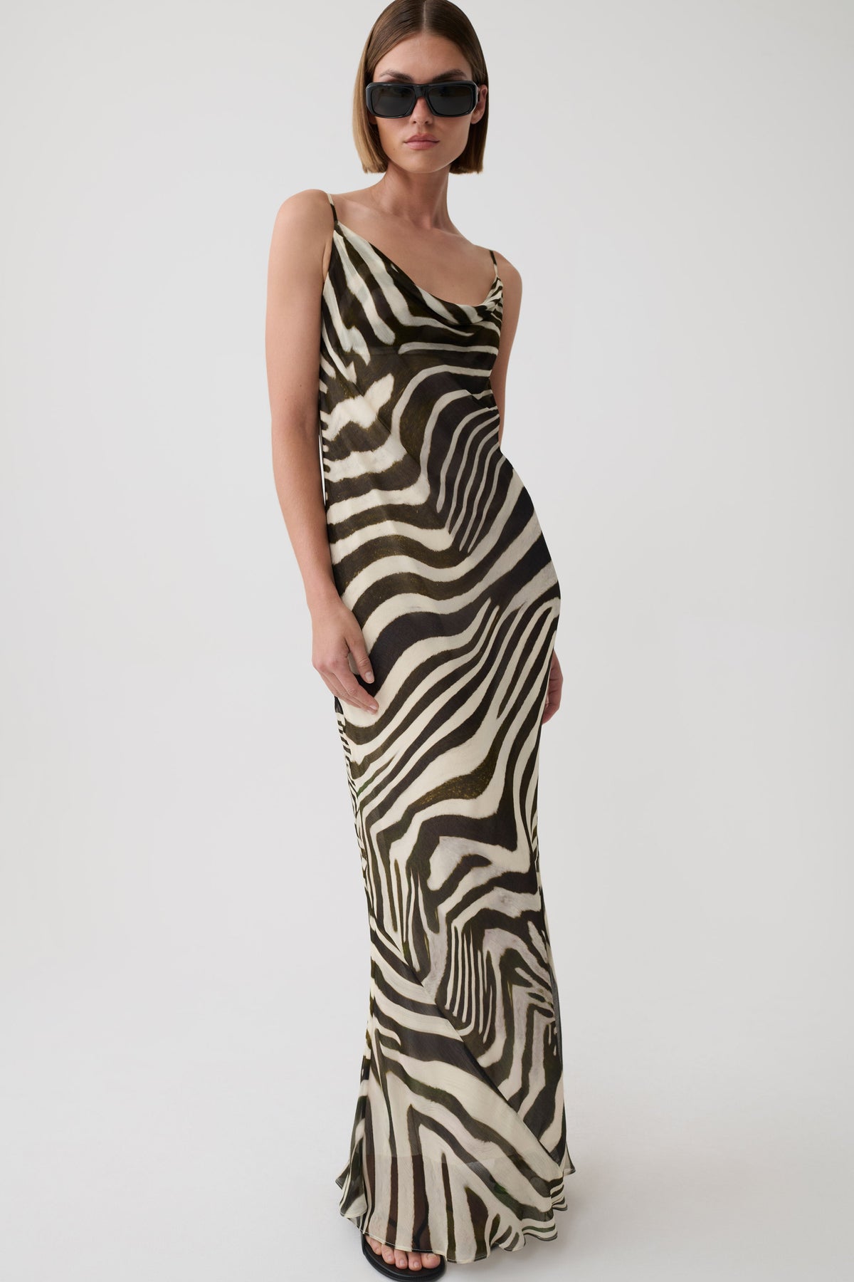 A woman modeling a sleeveless, floor-length zebra-print dress with a draped neckline and thin straps, photographed against a plain light gray studio background.