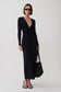 A woman modeling a long-sleeve black maxi dress with a deep V-neck and gathered waist, styled with black sunglasses, black strappy sandals, and a black woven handbag against a light gray studio background.