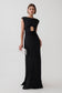 A woman modeling a floor-length black gown with a cutout at the waist and cap sleeves, holding a small white clutch against a plain light gray studio background.