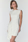 A woman modeling a sleeveless, off-white mini dress with a draped asymmetrical hem detail, set against a plain light gray studio background.
