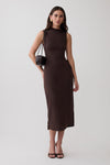 A woman modeling a sleeveless, high-neck, knee-length dark brown dress against a plain light gray studio background. She is carrying a small brown shoulder bag and wearing black heeled sandals.