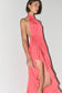 A woman modeling a coral pink halter-neck dress with a high-low ruffled hem and a side cutout, shown against a plain light gray studio background.