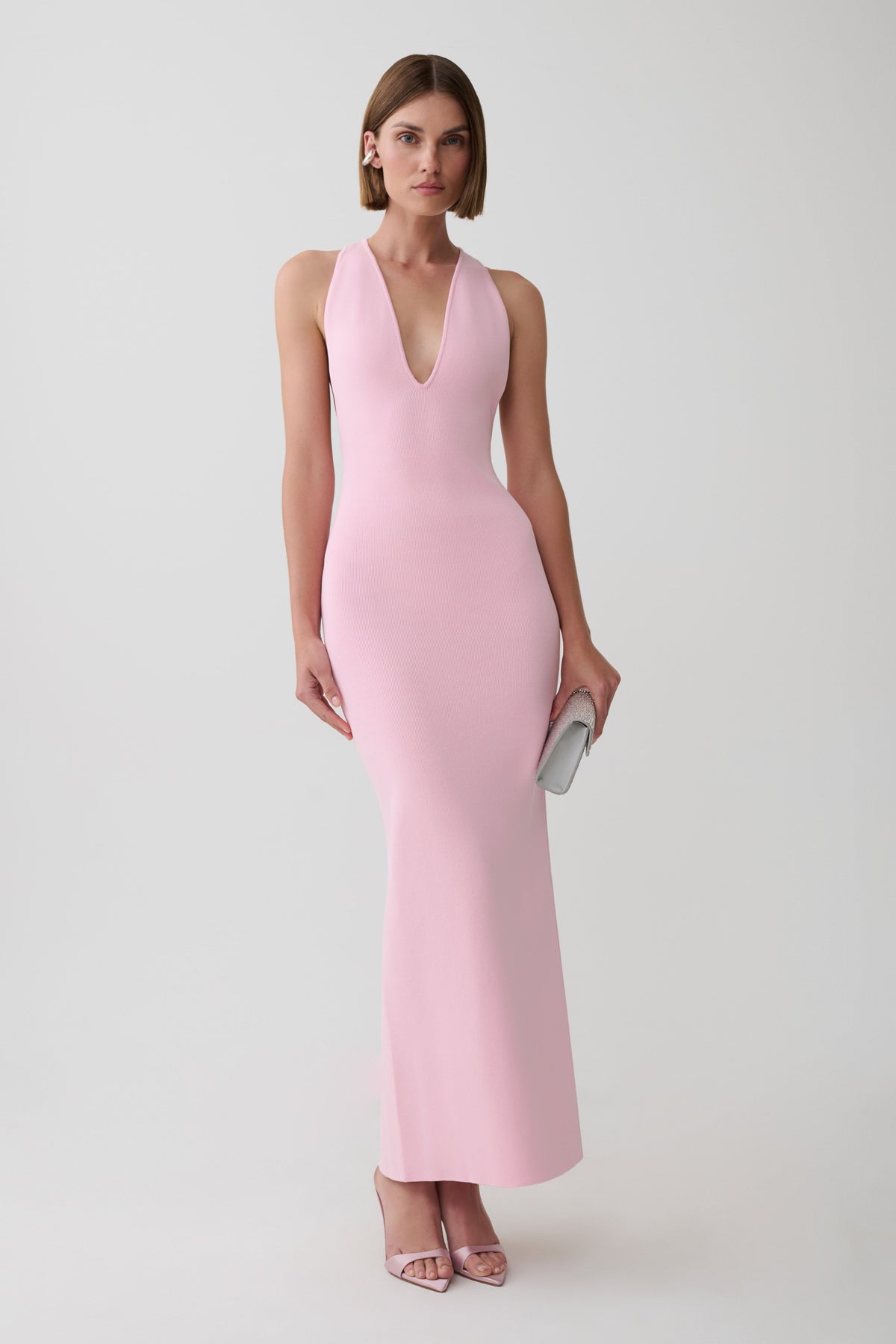 A woman modeling a sleeveless, light pink bodycon maxi dress with a deep V-neckline, standing against a plain light gray studio background.