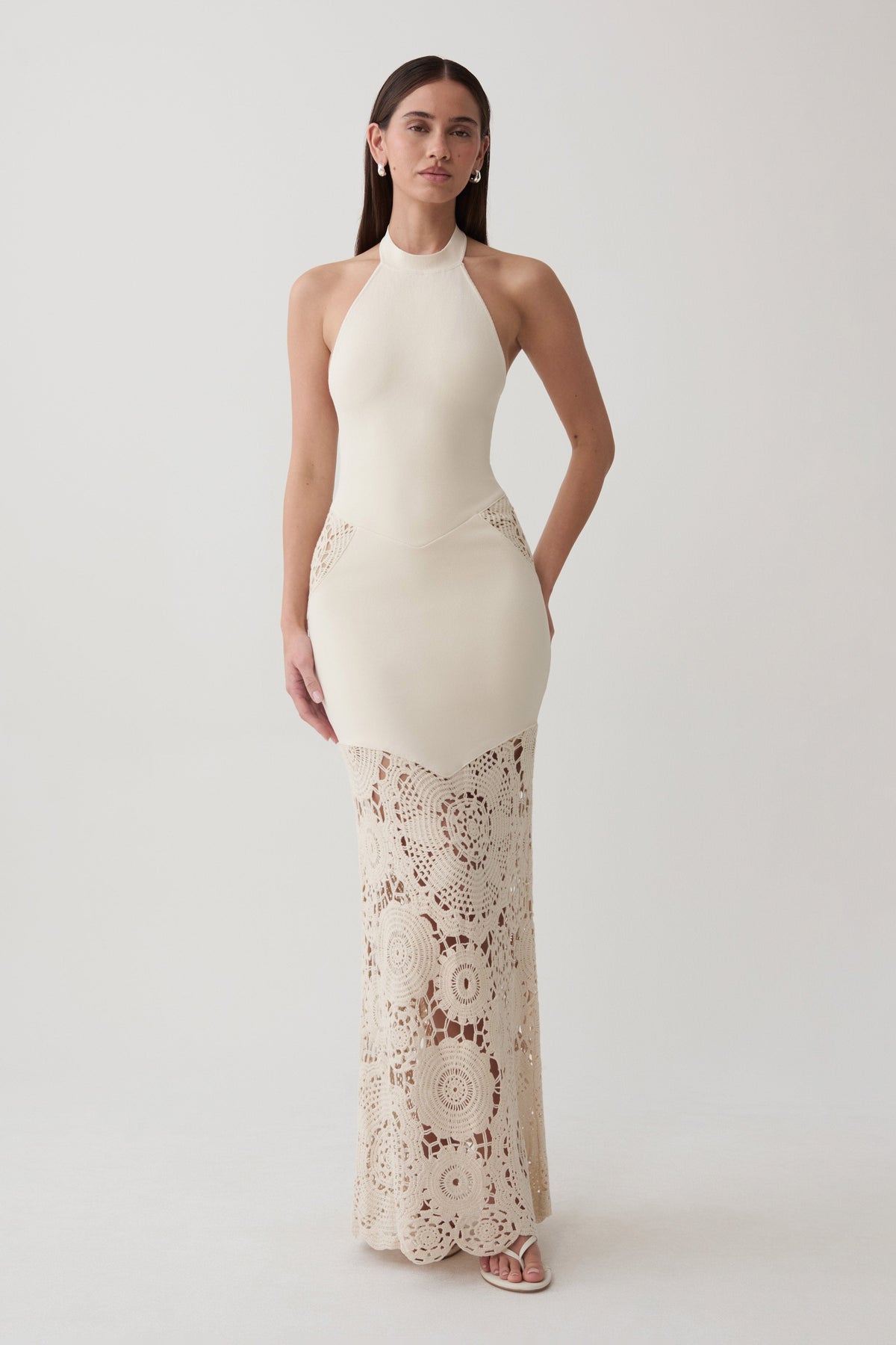 A woman modeling a sleeveless halter-neck maxi dress in cream with a textured crochet lace overlay on the lower half, set against a plain light gray studio background.