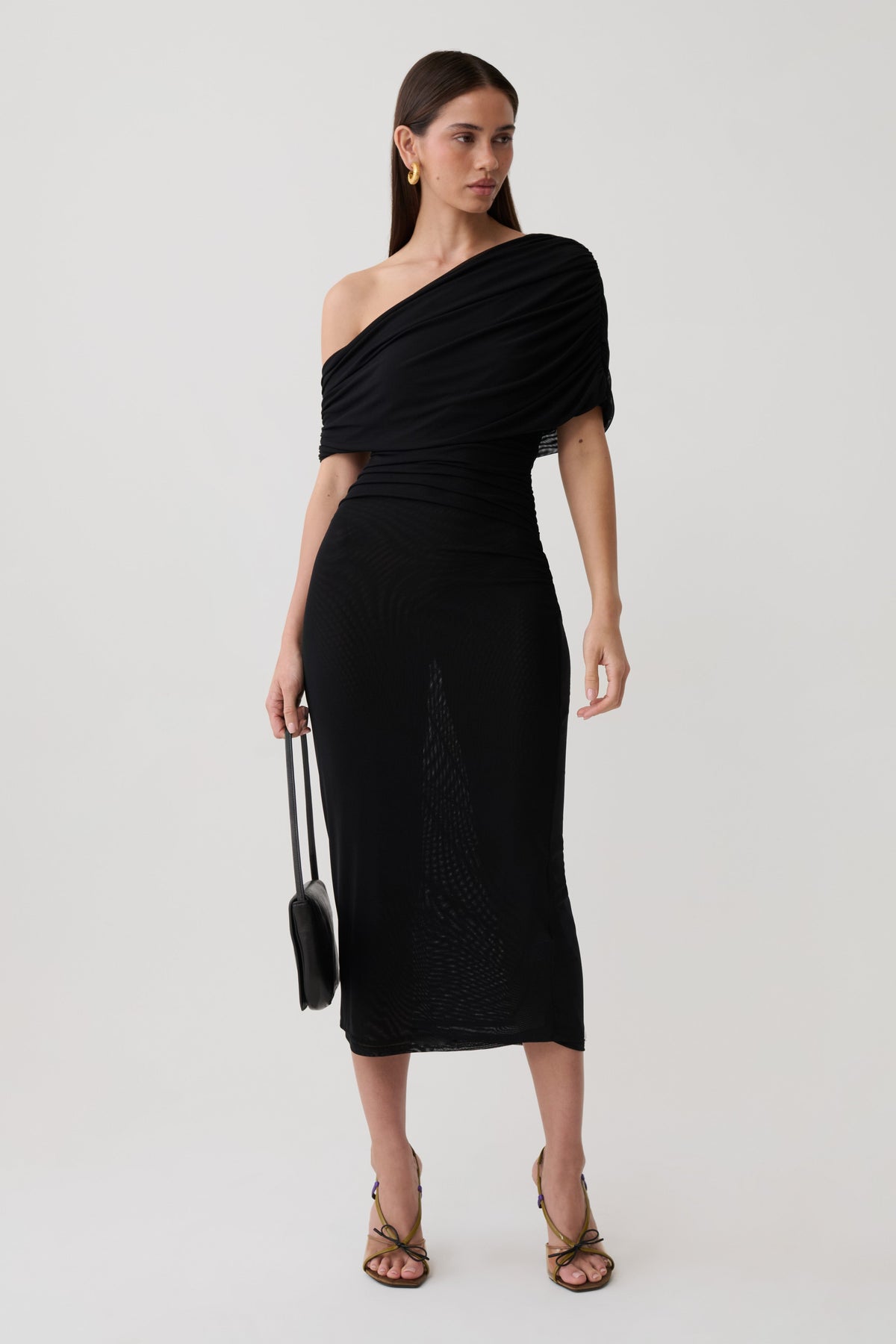 A woman modeling a black one-shoulder midi dress with textured knit detailing, holding a small black handbag against a light gray studio background.