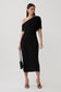 A woman modeling a black one-shoulder midi dress with textured knit detailing, holding a small black handbag against a light gray studio background.