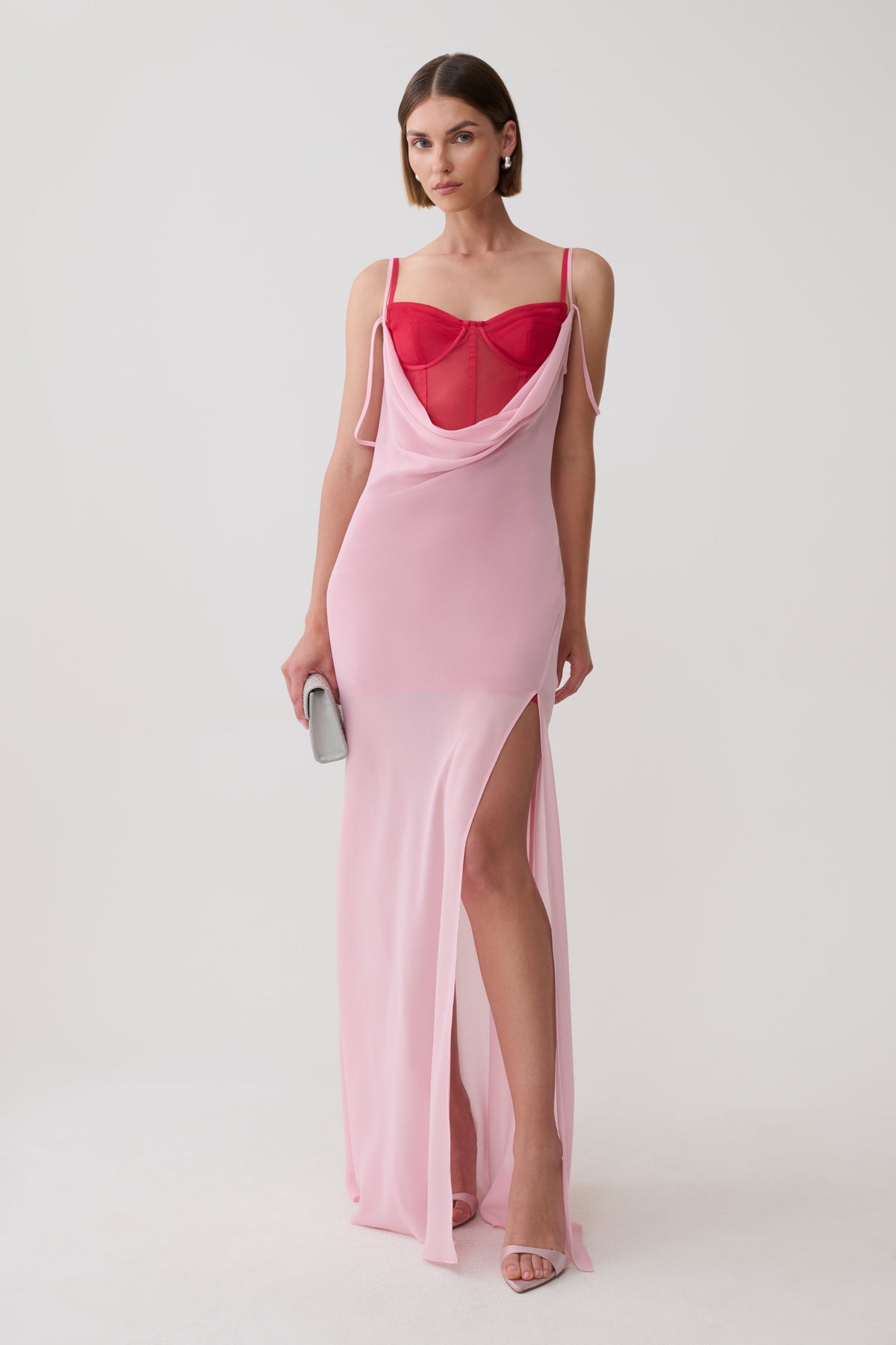 A model wearing a light pink slip-style maxi dress with a sweetheart neckline, sheer mesh bust, and a high thigh slit, paired with a bright red mesh corset mini dress underlayer and holding a small silver clutch against a plain light gray studio background.