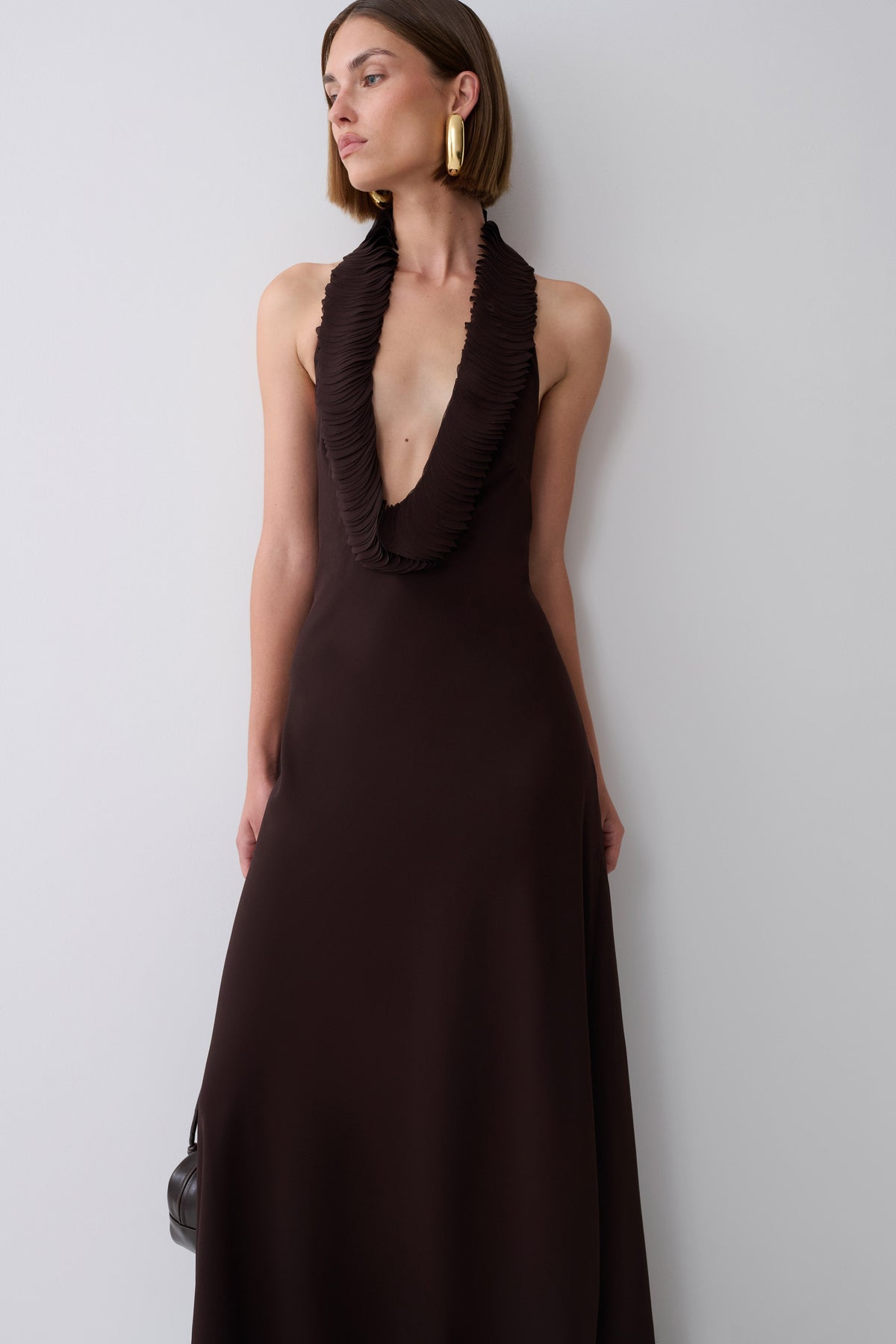 A woman wearing a sleeveless dark brown halter-neck maxi dress with a deep plunging ruffle neckline, standing against a light gray wall.