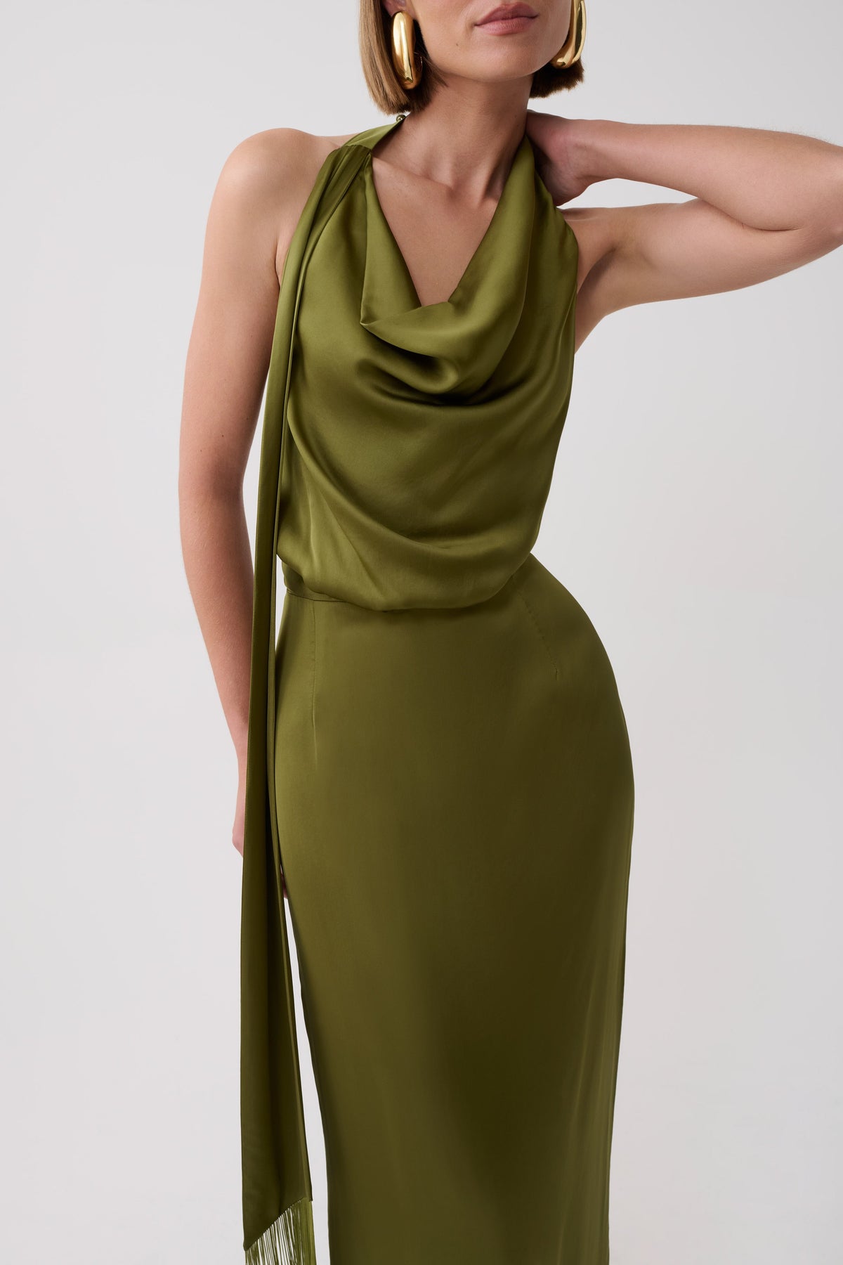 A woman wearing a draped olive green satin halter dress with a long flowing scarf detail, photographed against a clean light gray studio background.