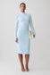 A model stands against a light gray studio background wearing a fitted light blue mock-neck midi dress with long sleeves, holding a small cream clutch and wearing clear-strap high-heel sandals.