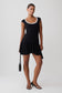 A woman modeling a black sleeveless mini dress with a white scalloped neckline, black strappy heels, and holding a black handbag against a light gray studio background.