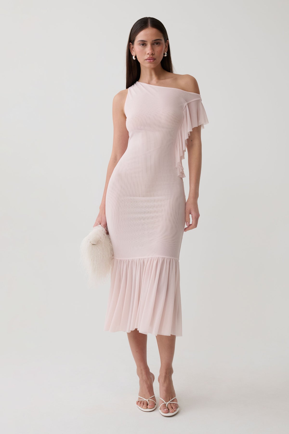 A woman models a light pink off-the-shoulder ribbed midi dress with a ruffled hem and side fringe detail, paired with white strappy heels and a white faux-fur clutch, against a plain light gray studio background.
