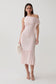 A woman models a light pink off-the-shoulder ribbed midi dress with a ruffled hem and side fringe detail, paired with white strappy heels and a white faux-fur clutch, against a plain light gray studio background.