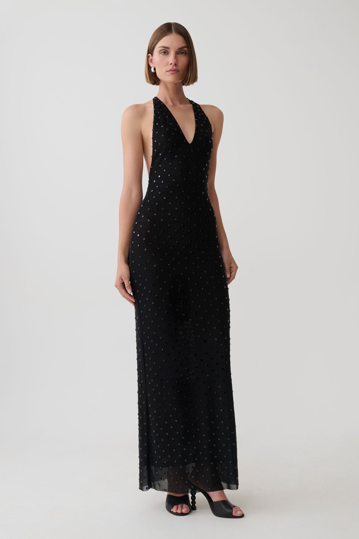 A woman modeling a sleeveless black halter-neck maxi dress with a subtle dotted pattern, photographed against a plain light gray studio background.