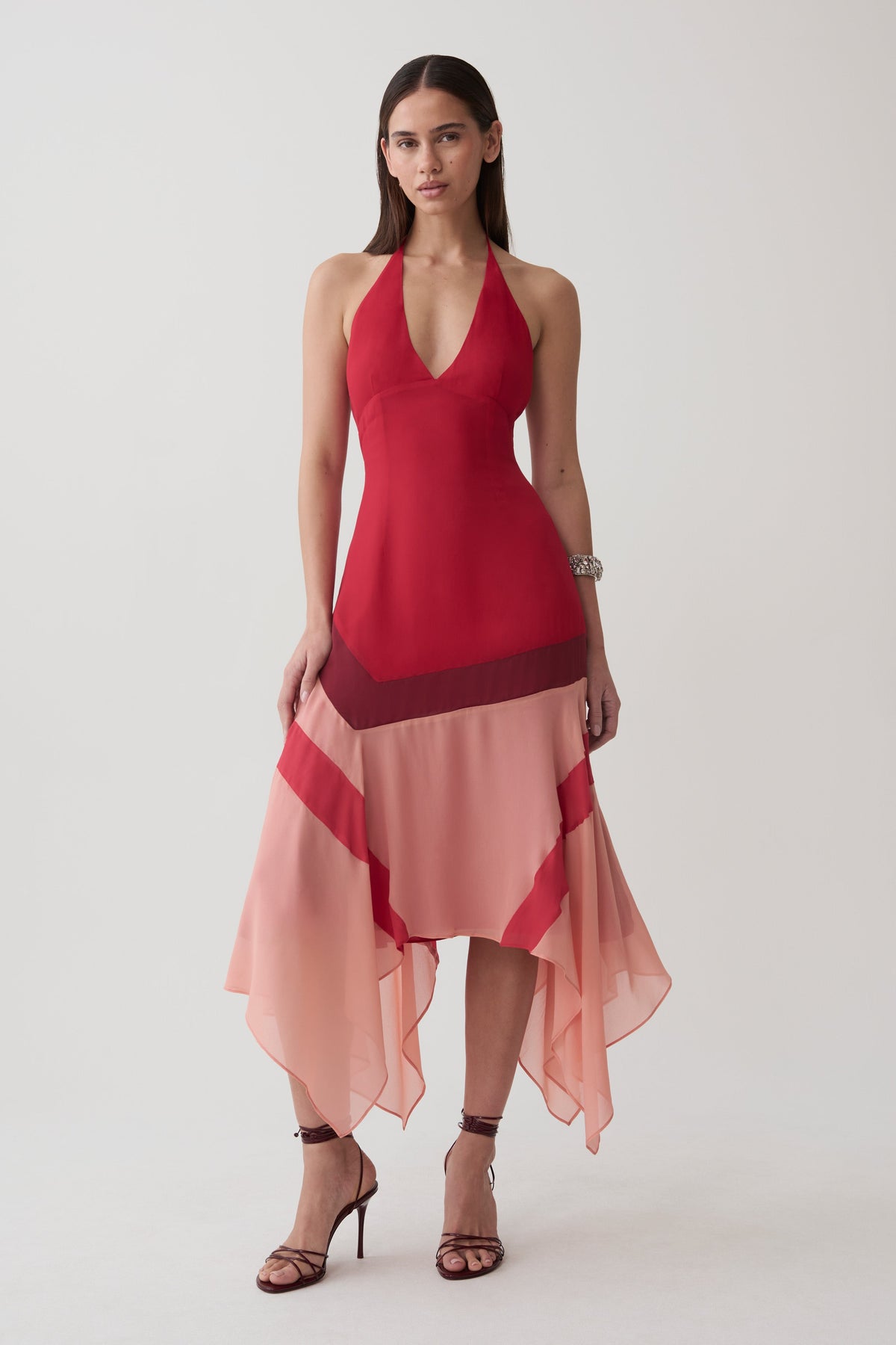 A woman modeling a sleeveless halter-neck midi dress with a deep V-neckline, featuring a solid red upper bodice and a layered, flowing peach-pink skirt with sheer side panels, set against a plain light gray studio background.