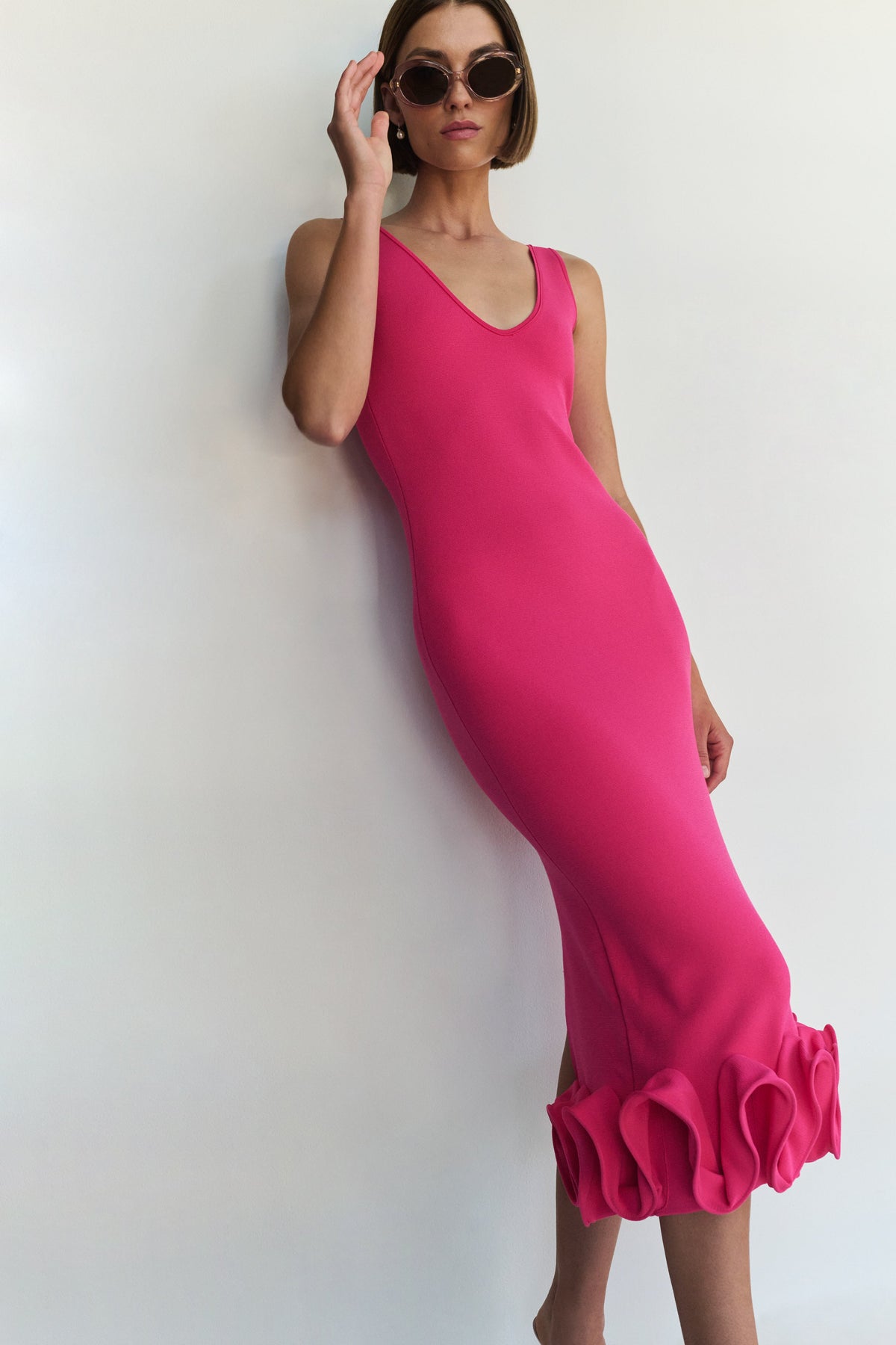 A woman wearing a bright pink sleeveless bodycon dress with a ruffled hem, posing against a plain white wall.