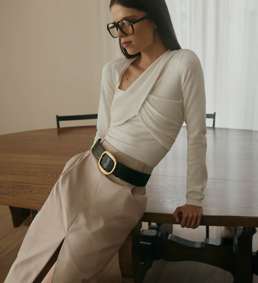 Woman sitting at a wooden table wearing a beige outfit with a black belt.