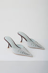 A pair of pointed-toe mule heels in a pale icy blue color, featuring decorative cutout detailing on the sides and slim dark brown stiletto heels, photographed on a clean white studio background.