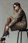 A model wearing a snakeskin-patterned long-sleeve jacket with matching midi skirt, black tights, and metallic snakeskin heels sitting on a black stool against a textured gray studio background.