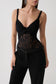 A model wearing a black corset-style camisole top with sheer lace paneling and black trousers, photographed against a plain light gray studio background.