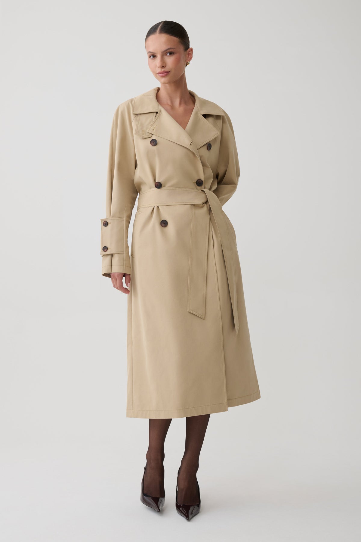 A woman modeling a mid-length beige trench coat with dark buttons, a matching waist belt, and dark tights with pointed heels, photographed against a plain light gray studio background.