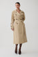 A woman modeling a mid-length beige trench coat with dark buttons, a matching waist belt, and dark tights with pointed heels, photographed against a plain light gray studio background.
