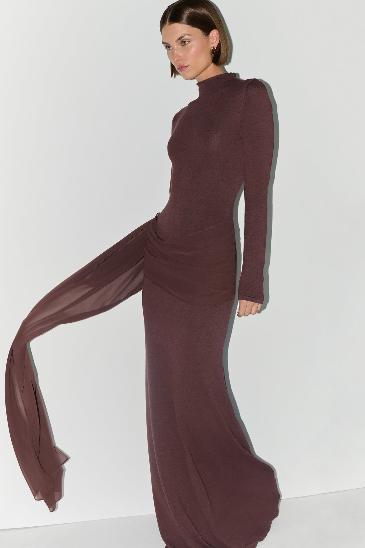 A woman modeling a long-sleeve, high-neck, floor-length dress in a deep plum-brown color, featuring a draped sheer side panel that flows out to the side against a plain white studio background.
