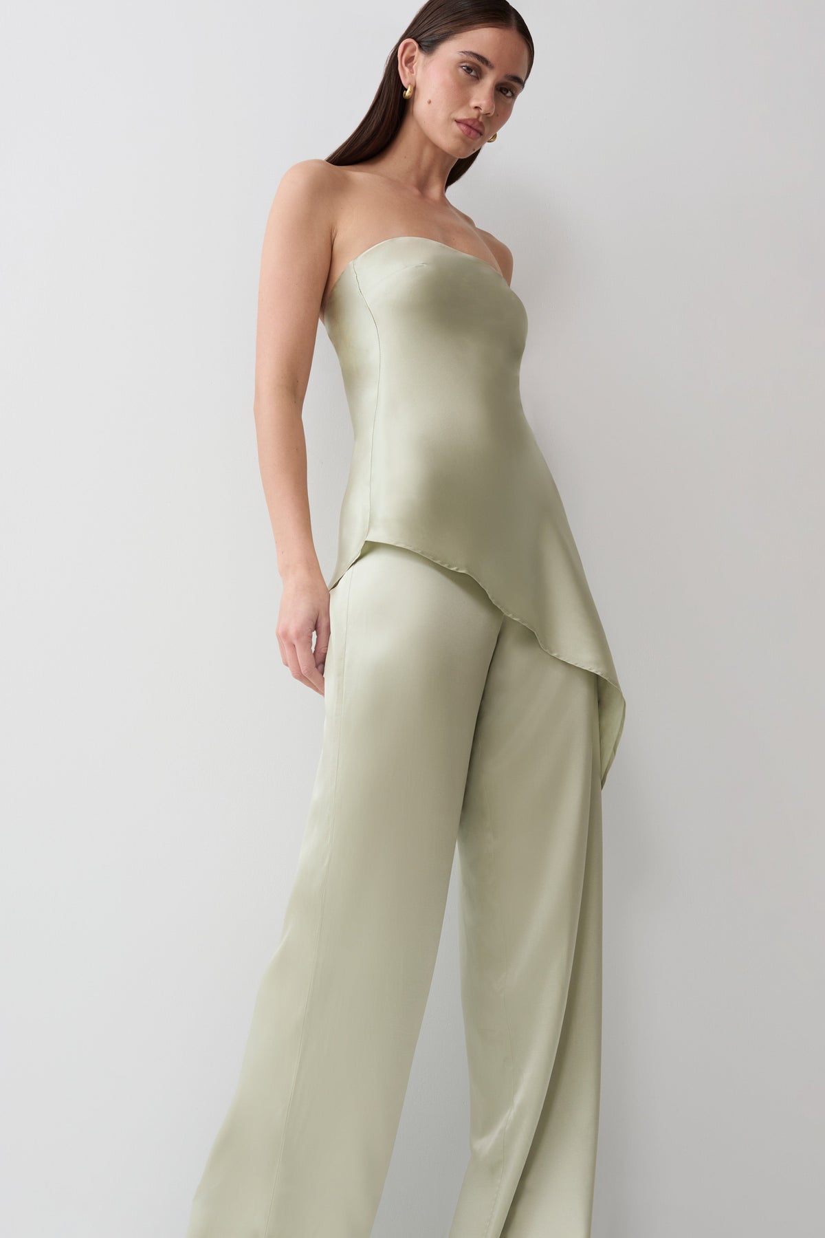 A woman modeling a strapless satin-like jumpsuit in a light sage green color, set against a plain light gray studio background.
