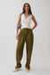 A model stands against a light gray studio background wearing olive green wide-leg trousers, a white sleeveless top, and cream pointed-toe heels, while carrying a black-and-white patterned shoulder bag.