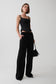 A model stands against a light gray studio background wearing a black sleeveless top, black wide-leg trousers, and holding a small black feather-trim handbag.
