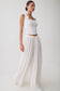 A woman modeling a white sleeveless crop top with scalloped trim and matching white wide-leg pleated pants against a plain light gray studio background.