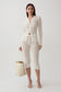 A model stands against a light gray studio background wearing a matching cream lace three-piece set consisting of a long-sleeve button-front cardigan, cropped capri pants, and a tie belt. She is holding a cream canvas tote bag and wearing strappy high-heeled sandals.