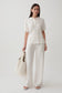A woman modeling a matching two-piece outfit consisting of a short-sleeve collared blouse and wide-leg trousers in an off-white lace fabric, holding a fluffy cream tote bag against a plain light gray studio background.
