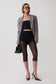 A model wearing a gray blazer, black crop top, black high-waisted shorts, sheer black capri tights, black pointed-toe heels, and holding a black handbag against a light gray studio background.