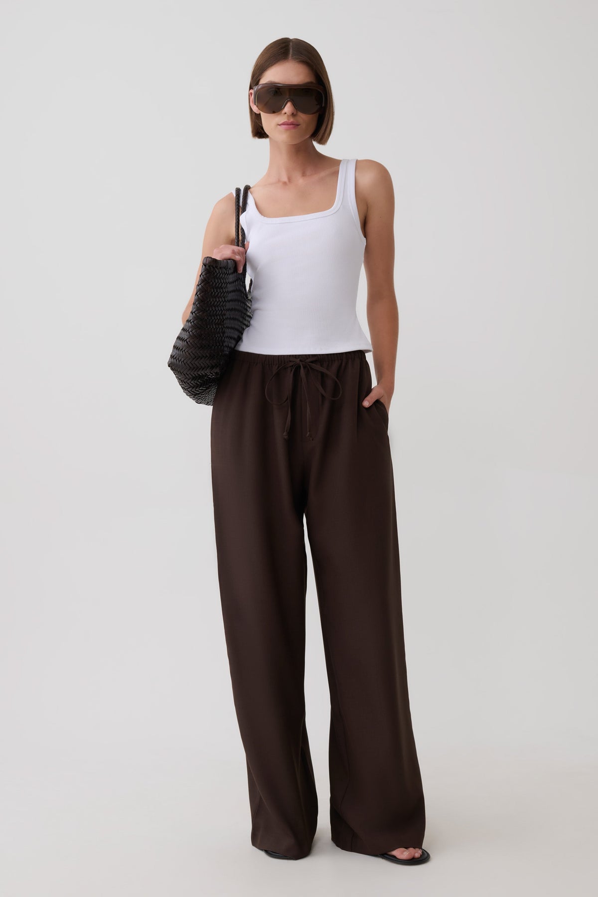 A model wearing a white tank top, dark brown wide-leg drawstring pants, black sunglasses, and carrying a black textured shoulder bag against a light gray studio background.