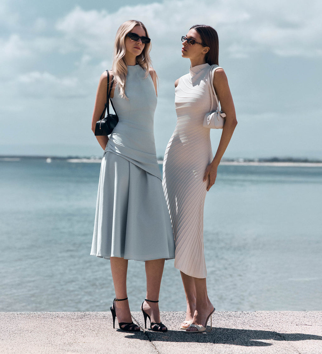 Two women at the pier. One in blue high neck midi dress, the other in a while high neck midi dress. 