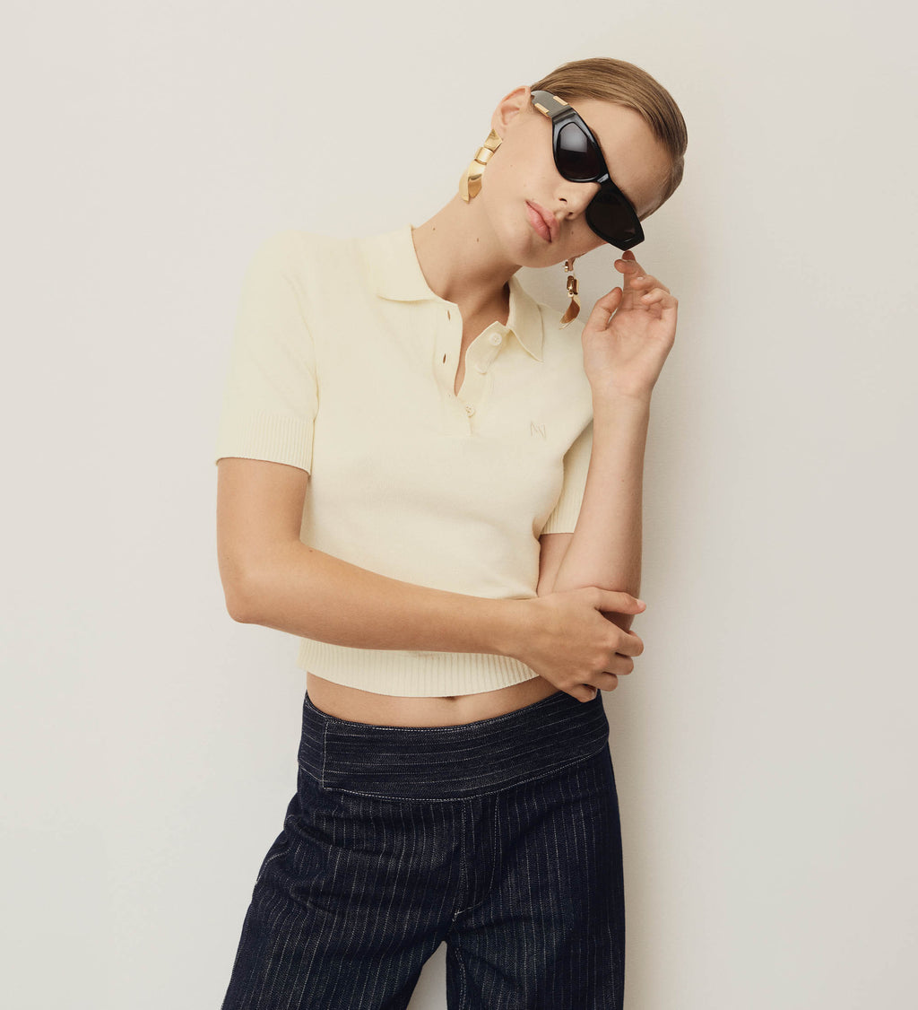 Woman wearing a beige cropped top and dark pinstripe pants with sunglasses on a plain background.