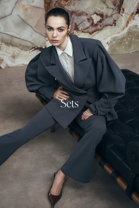 Woman in a dark gray suit sitting on a couch with a textured wall background.