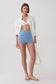A model stands against a light gray studio background wearing a white open-front shirt, a white bralette, light blue high-waisted shorts, and white strappy sandals, while holding a white woven tote bag.