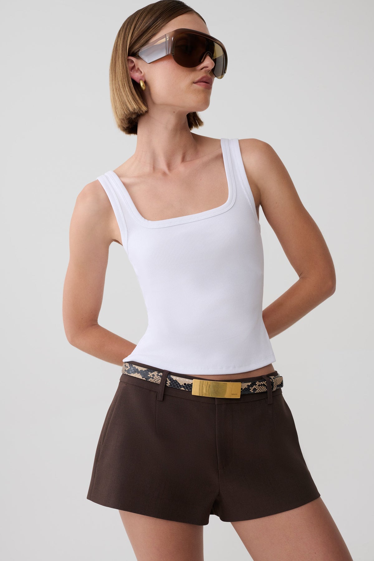 A model wearing a white scoop-neck tank top and dark brown shorts with a patterned belt, styled with oversized sunglasses against a light gray background.