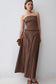 A woman models a strapless brown maxi top and skirt set with frayed trim details, holding a small leopard-print clutch against a plain light gray wall.