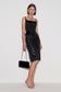 A model wearing a black satin camisole top and a black studded pencil skirt, holding a black velvet clutch bag, posed against a plain white studio background.