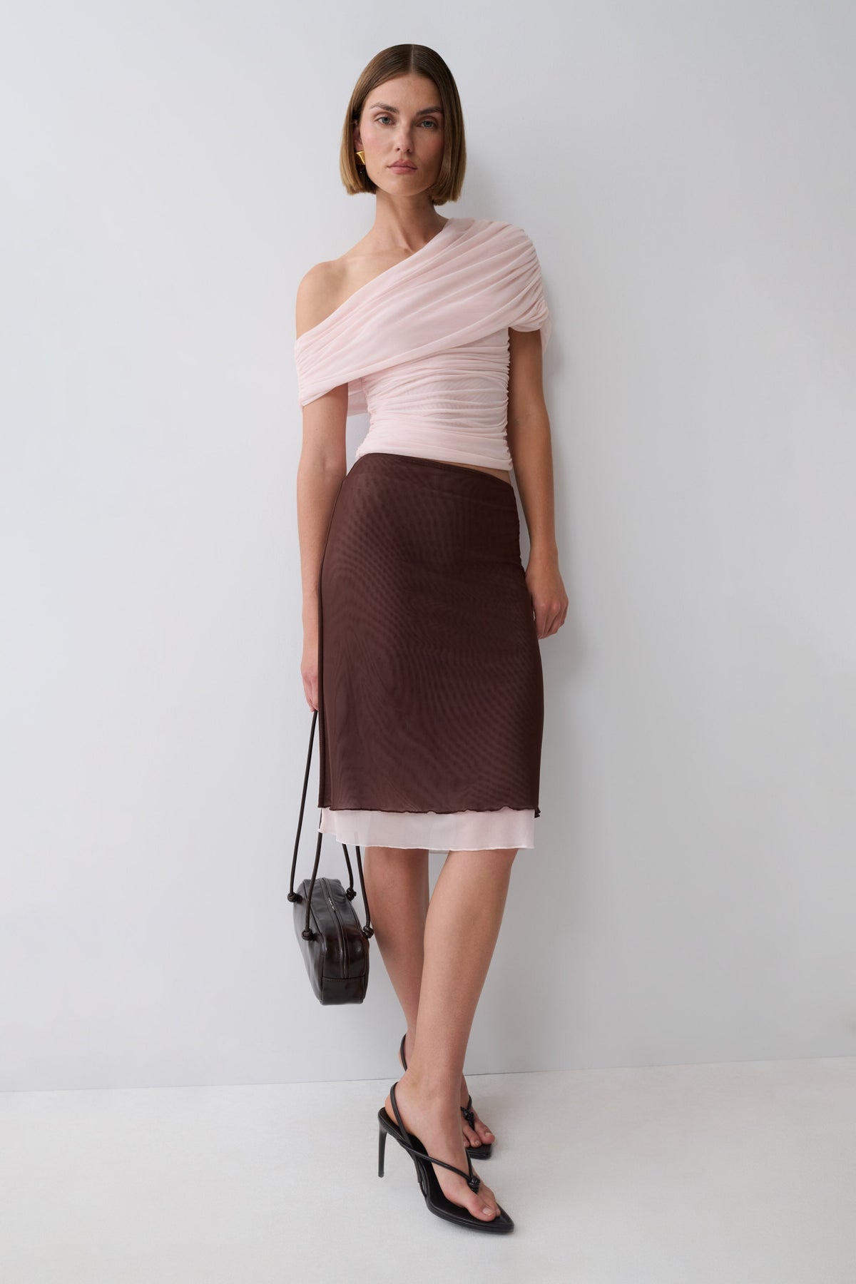 A model stands against a light gray studio wall wearing a pale pink one-shoulder top and a dark brown textured pencil skirt with a sheer pink underlayer. She is holding a small black handbag and wearing black strappy heels.