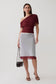 A model stands against a light gray studio background wearing a draped off-the-shoulder burgundy top and a fitted white pencil skirt with a sheer burgundy hem, holding a small black handbag.