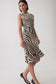 A woman modeling a sleeveless halter-neck midi dress with a bold black and beige zebra-like wavy stripe pattern, paired with black heels, against a plain light gray studio background.