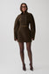 A model wearing a dark brown double-breasted mini coat with oversized sleeves, paired with sheer black tights and dark pointed-toe heels, standing against a light gray studio background.