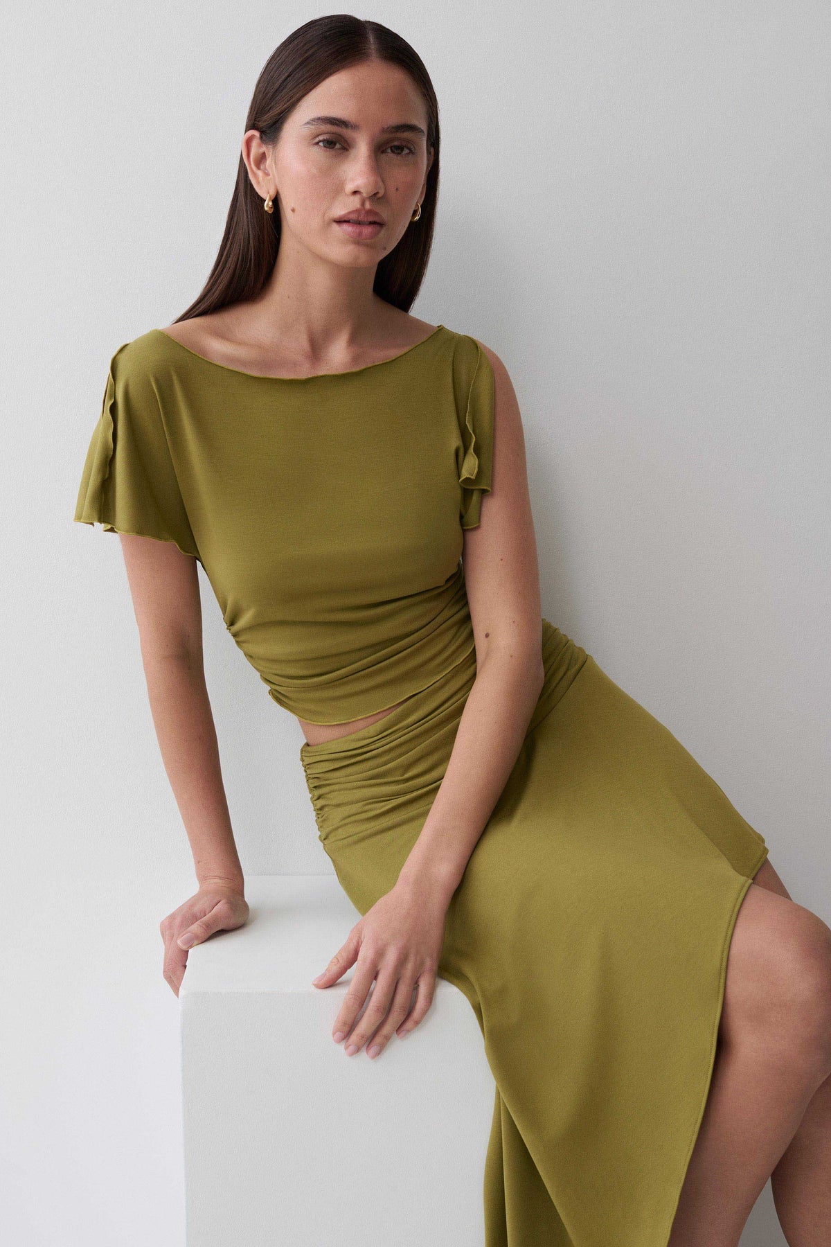 A woman wearing an olive green midi dress with flutter sleeves and a ruched bodice, leaning against a white block in a minimalist studio setting.